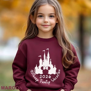 A young girl with long blonde hair wearing a maroon sweater with a white Disney castle and text printed on it.