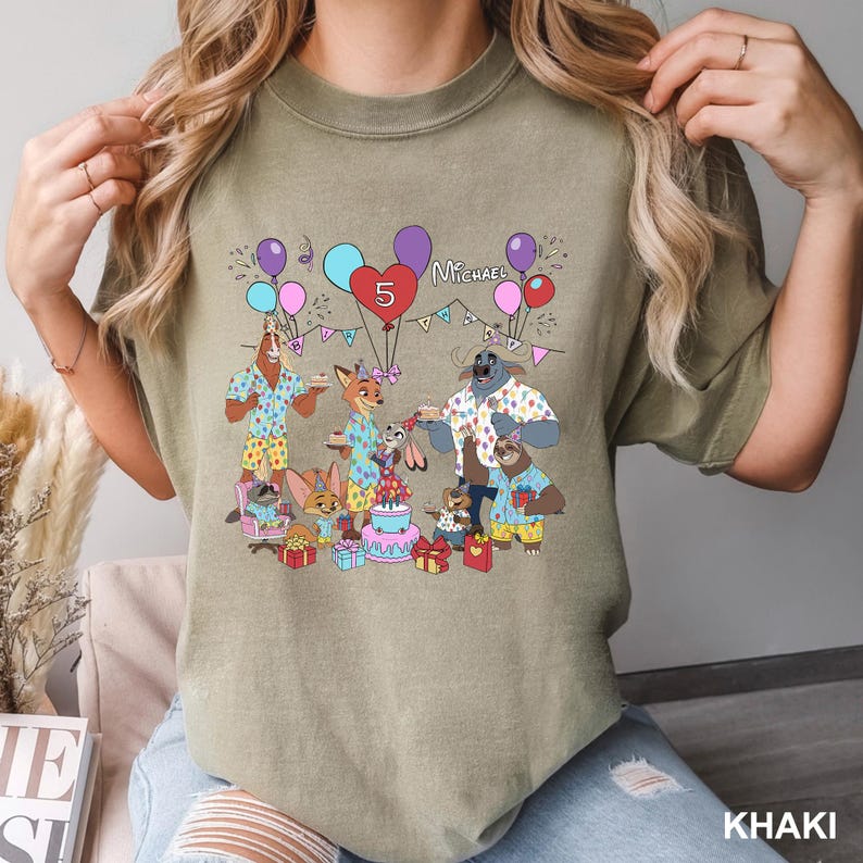 A person wearing a t-shirt with a cartoon illustration of a birthday party scene featuring a dog, cat, and other animals.