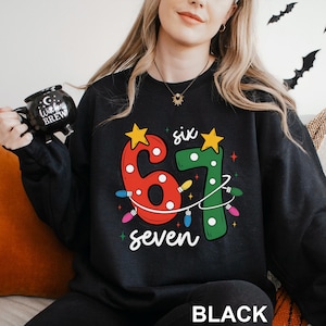 May include: Black sweatshirt with a festive "67" design in red and green, adorned with string lights and stars. The word "six" is above the "6" and "seven" is below the "7". The word "BLACK" is at the bottom.