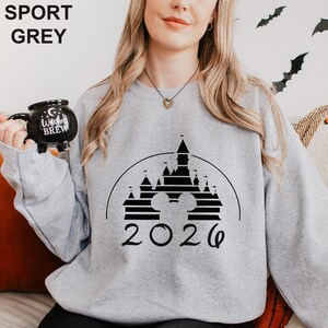 A woman wearing a grey sweatshirt with a black and white graphic of a castle and the year "2026" printed on it. She is holding a black mug with the text "I love Brew" printed on it.