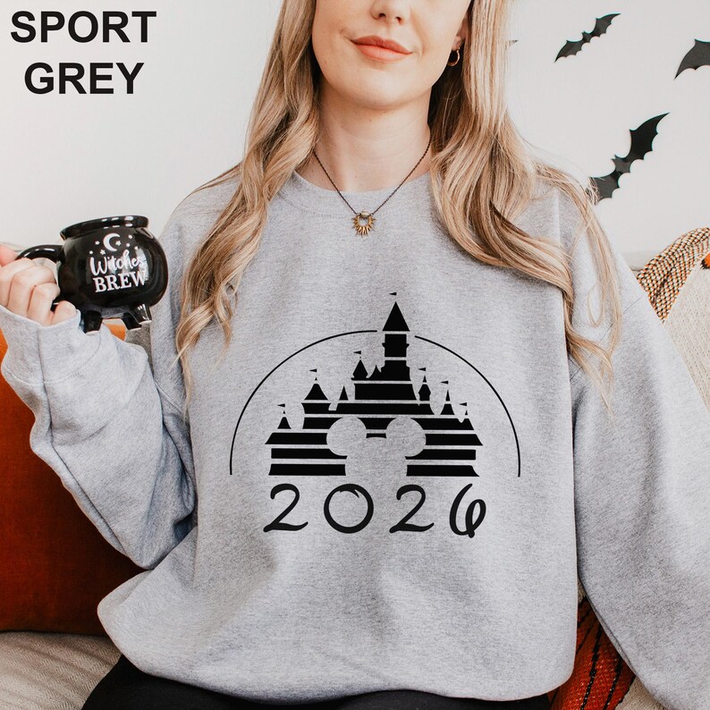 A woman wearing a grey sweatshirt with a black and white graphic of a castle and the year "2026" printed on it. She is holding a black mug with the text "I love Brew" printed on it.