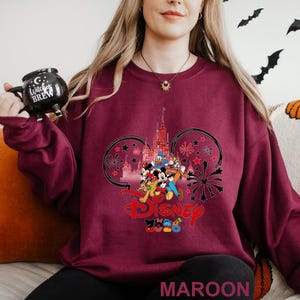 A woman wearing a purple sweater with a Disney logo and a mug with the text "C witch brew" is sitting on a couch.