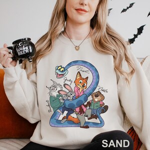 A woman wearing a white sweatshirt with a cartoon character design, holding a mug with a black and white design that says "Witch's Brew".