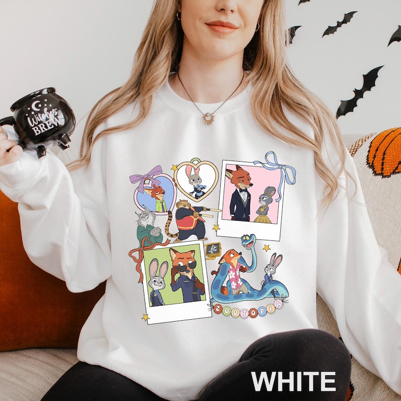 A woman wearing a white sweatshirt with a collage of cartoon animal characters on it, including a fox, rabbit, and other animals. She is holding a mug with a black design and is sitting on a couch.