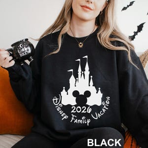 A woman wearing a black sweatshirt with a white castle and Mickey Mouse design, holding a black mug with the text "Disney 2020 Family Vacation".