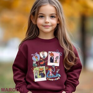 A young girl with long blonde hair is wearing a maroon sweater with a collage of cartoon animal characters on it.