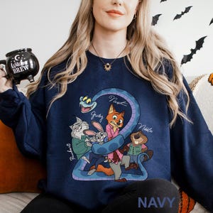 A woman wearing a navy blue sweatshirt with a cartoon character design, including a cat, rabbit, and other animals, is sitting on a couch. She is holding a black mug with a Halloween-themed design.