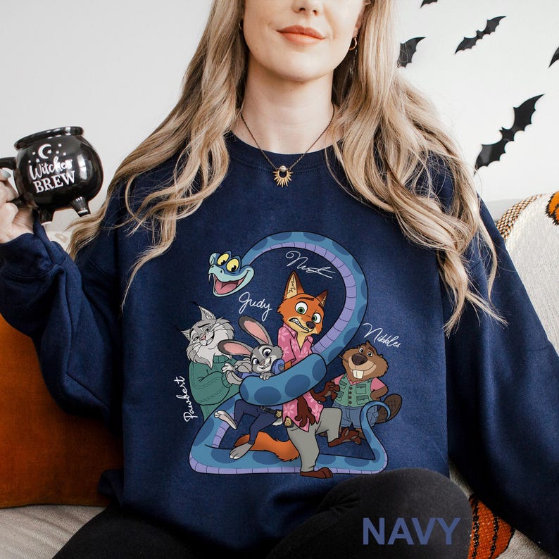 A woman wearing a navy blue sweatshirt with a cartoon character design, including a cat, rabbit, and other animals, is sitting on a couch. She is holding a black mug with a Halloween-themed design.