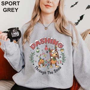 A woman wearing a grey sweatshirt with a cartoon dog and the text "DASHING Through The Show" printed on it. She is holding a black mug with the text "I love Brew" printed on it.
