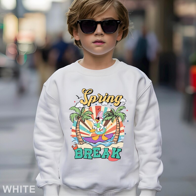 A young boy wearing a white sweatshirt with a colorful graphic design and sunglasses stands in an urban setting.