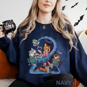 A woman wearing a navy blue sweatshirt with a cartoon character design, including a cat, rabbit, and other animals, is sitting on a couch. She is holding a black mug with a Halloween-themed design.