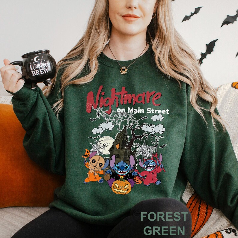 A woman wearing a green sweatshirt with a Halloween-themed design, including characters from the movie "Nightmare on Main Street". She is holding a black mug with a Halloween-themed design.