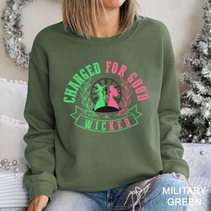May include: Military green sweatshirt with the text "CHANGED FOR GOOD" and "WICKED" in a circular design. The design features silhouettes of two figures, one green and one pink, with a clock face in the background. The sweatshirt is a crew neck style.