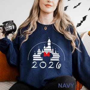 A woman wearing a navy blue sweater with a white castle graphic and the year "2026" printed on it, holding a black mug with a white design.