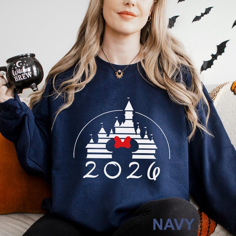 A woman wearing a navy blue sweater with a white castle graphic and the year "2026" printed on it, holding a black mug with a white design.