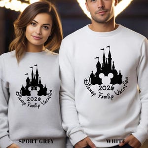 A woman and a man standing next to each other, both wearing white sweatshirts with a black silhouette of a castle and the text "Disney Family Vacation" printed on them.