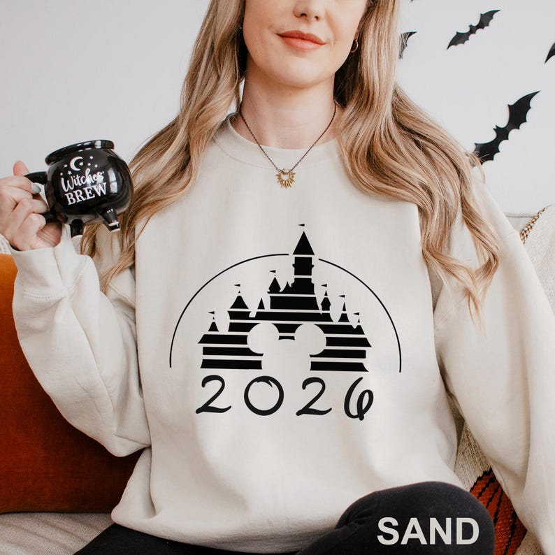 A woman wearing a white sweater with a black castle graphic and the year "2026" printed on it, holding a mug with a "Witch's Brew" design.
