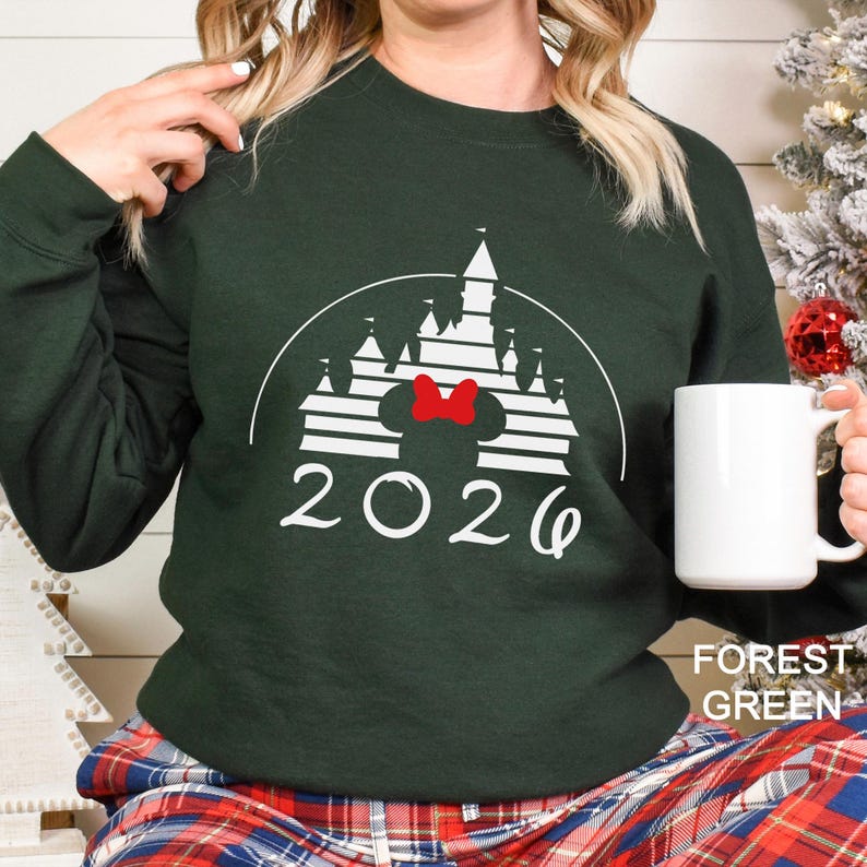 A woman wearing a green sweater with a white castle graphic and the year "2026" printed on it, holding a white mug.