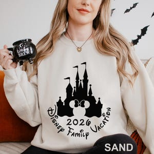 A woman wearing a white sweater with a black castle and Minnie Mouse design, holding a black mug with the text "Witch's Brew" and "Disney Family Vacation" printed on it.