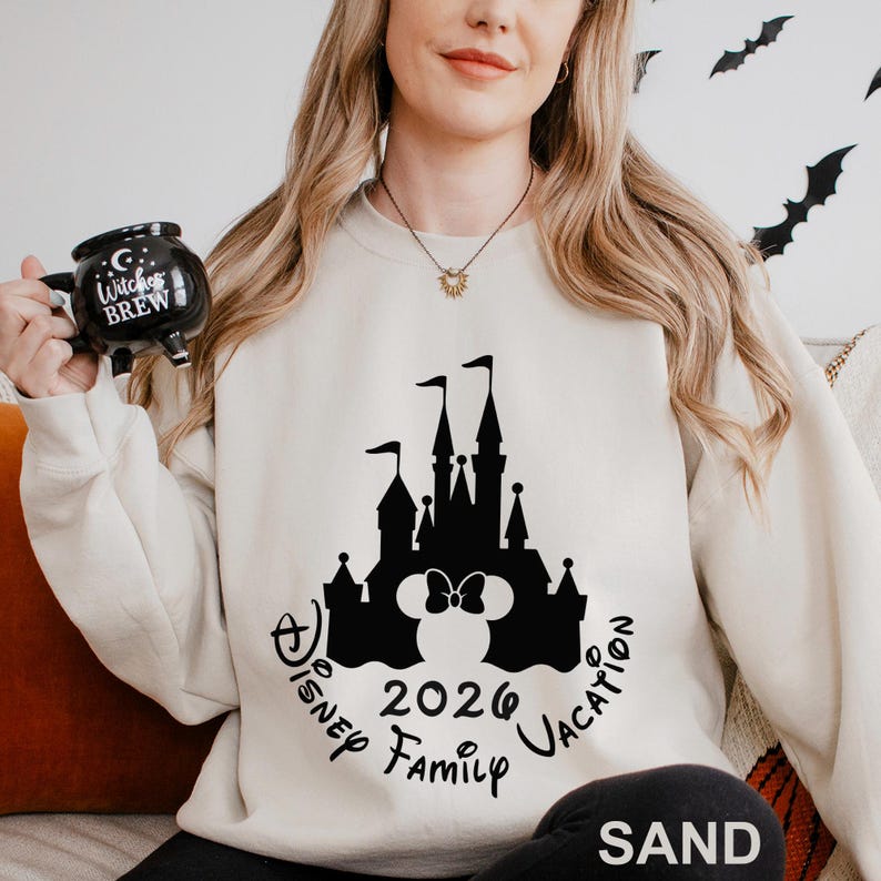 A woman wearing a white sweater with a black castle and Minnie Mouse design, holding a black mug with the text "Witch's Brew" and "Disney Family Vacation" printed on it.