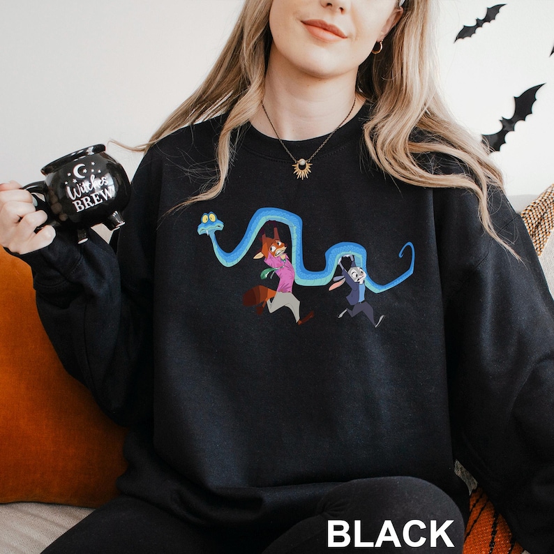 A woman wearing a black sweater with a cartoon character design, holding a black mug with the text "Witch Brew" printed on it.