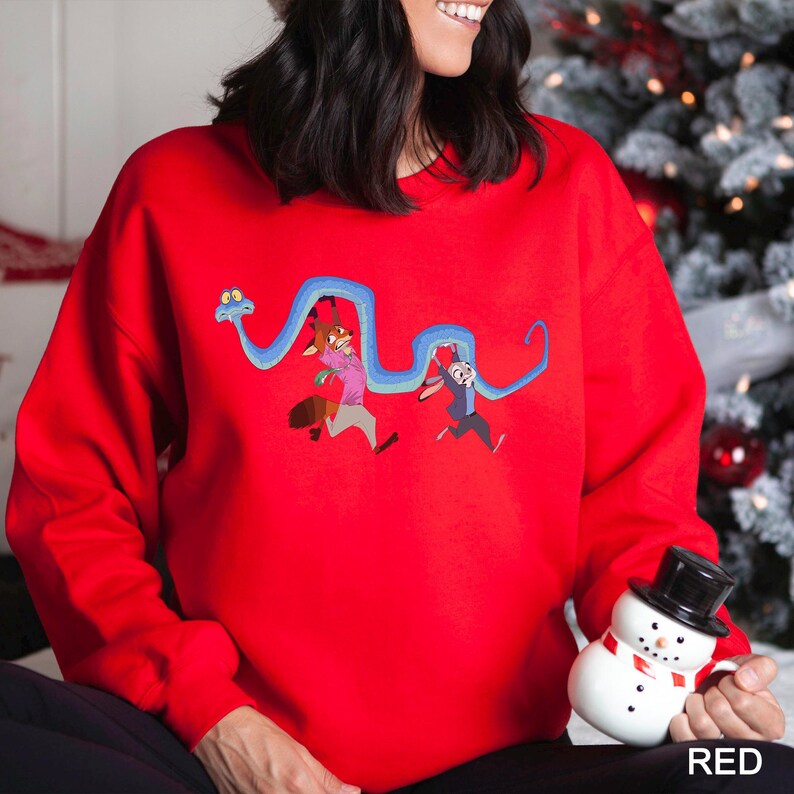 A woman wearing a red sweater with a cartoon character design is holding a snowman and a mug.
