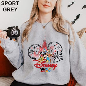 A woman wearing a grey sweatshirt with a Disney-themed design, including Mickey Mouse and Minnie Mouse characters, and a castle in the background. She is holding a black mug with the text "Disney BREW" printed on it.