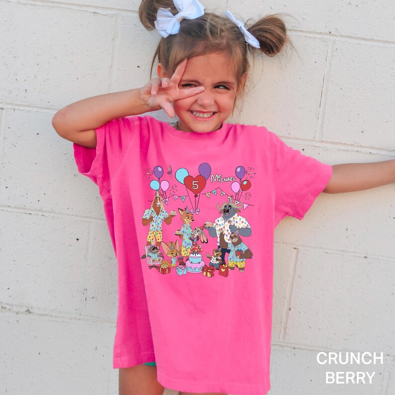 A young girl wearing a pink t-shirt with a cartoon character design and balloons, smiling and making a peace sign with her fingers.