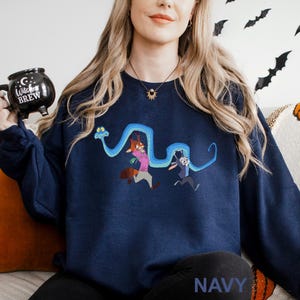 A woman wearing a navy blue sweatshirt with a cartoon character design, holding a black mug with a "Witch Brew" label.