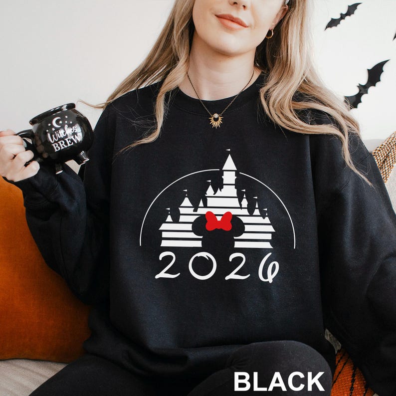 A woman wearing a black sweatshirt with a white and red Disney castle graphic and the year "2026" printed on it, holding a black mug with the text "Witch Brew" printed on it.