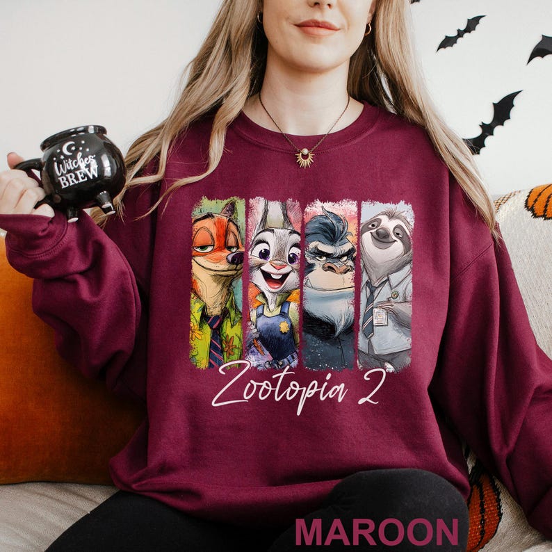A woman wearing a burgundy sweatshirt with a graphic of the four animated characters from the movie "Zootopia" on it, holding a black mug with the text "COOK BREW" printed on it.