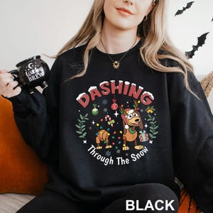 A woman wearing a black sweatshirt with a Christmas-themed design, including a cartoon dog and snowflakes, holding a black mug with the text "Witch Brew" printed on it.