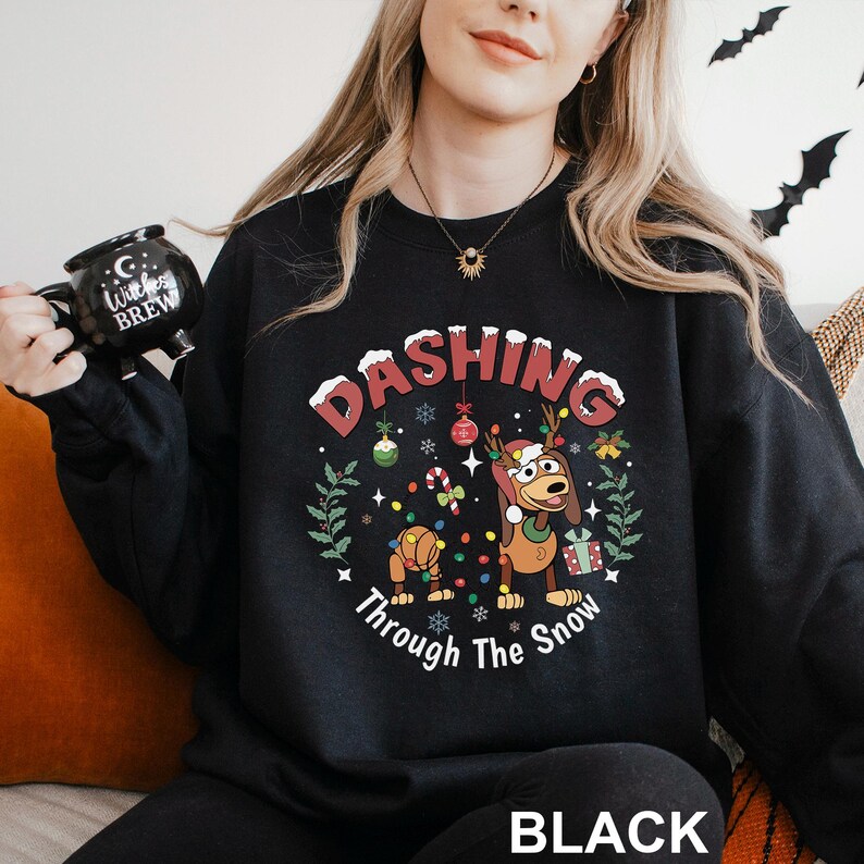 A woman wearing a black sweatshirt with a Christmas-themed design, including a cartoon dog and snowflakes, holding a black mug with the text "Witch Brew" printed on it.