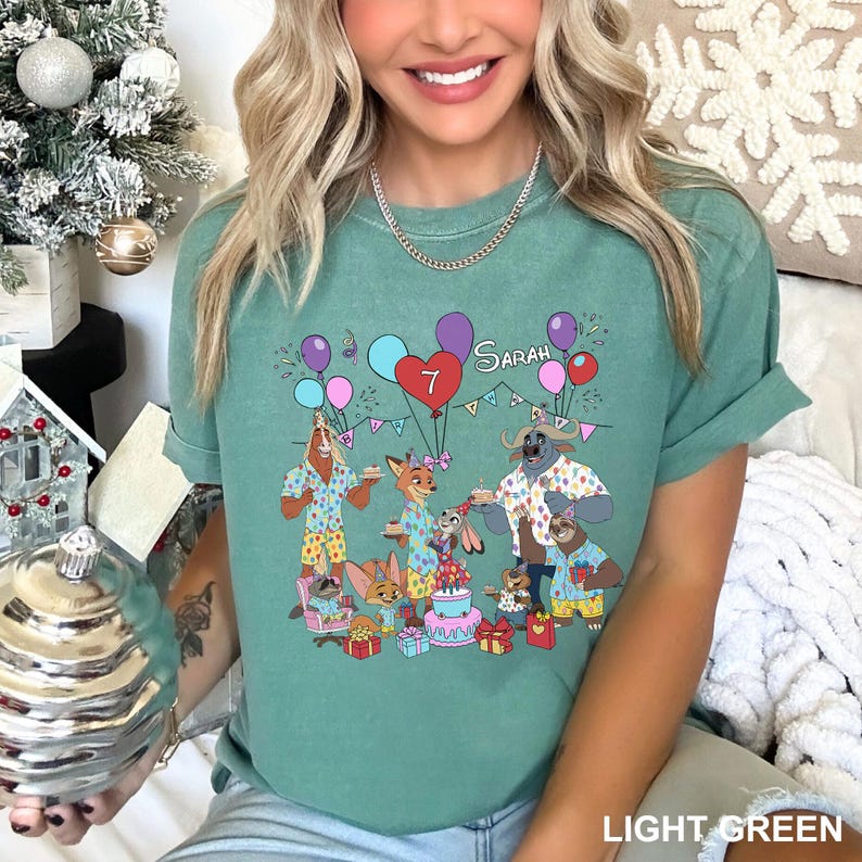 A woman wearing a green t-shirt with a cartoon illustration of three animals, including a dog, a cat, and a bird, surrounded by balloons and presents, with the text "Sara" on the shirt.