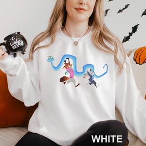 A woman wearing a white sweater with a cartoon character design is sitting on a couch. She is holding a mug with a black design and a blue snake.