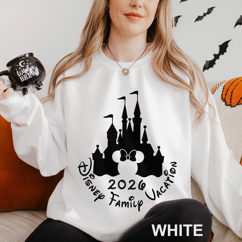 A woman wearing a white sweater with a black silhouette of a castle and the text "Disney Family Vacation" printed on it, holding a mug with a black design.