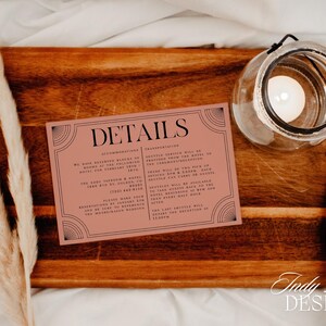 May include: A wedding details card with the word "DETAILS" in black lettering, set on a wooden tray with a candle in a glass jar. The card has a pink background and black decorative borders. Dried pampas grass adds a natural touch.