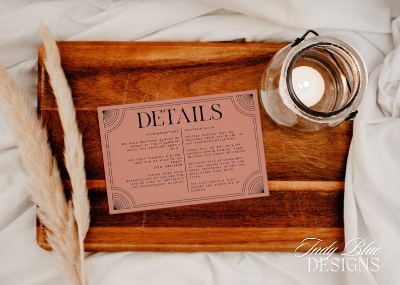 May include: A wedding details card with the word "DETAILS" in black lettering, set on a wooden tray with a candle in a glass jar. The card has a pink background and black decorative borders. Dried pampas grass adds a natural touch.