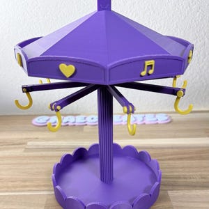 May include: A purple carousel-style jewelry stand featuring a central fluted pillar, a scalloped circular base, and a rotating top canopy. The canopy is decorated with yellow heart and musical note icons, and includes yellow hooks for hanging items.