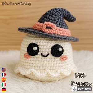Puede incluir: Un fantasma tejido a ganchillo con un sombrero de bruja. El fantasma es de color crema con ojos negros, una pequeña sonrisa y mejillas rosadas. El sombrero es gris con una banda y hebilla rosas. Incluye el texto "PDF Pattern Download".