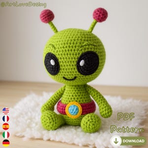 Puede incluir: Una muñeca alienígena verde tejida a ganchillo con grandes ojos negros, una boca sonriente y puntas de antena rosas. Tiene un cinturón rojo y dorado. La muñeca está sentada sobre una superficie blanca y esponjosa. Incluye el texto "PDF Pattern".