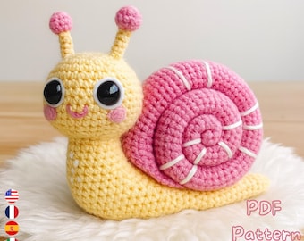 May include: A handmade crochet snail toy in yellow and pink. The snail has large black eyes, pink cheeks, and a smiling mouth. The shell is a spiral of pink with white stripes. The text "PDF Pattern" and a download arrow are visible.