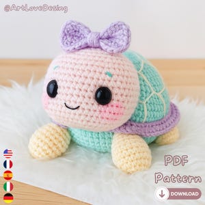 Peut inclure: Une tortue crochetée aux couleurs pastel, avec une tête rose clair ornée d'un nœud violet, des yeux noirs et des joues roses. La carapace est turquoise et violette. Le texte "PDF Pattern" et "Download" est inclus.