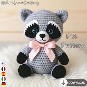 Puede incluir: Un peluche de mapache tejido a ganchillo con cuerpo gris, marcas faciales en blanco y negro y un lazo rosa. El mapache tiene ojos azules y patas negras. La imagen incluye el texto "PDF Pattern" y un icono de descarga.