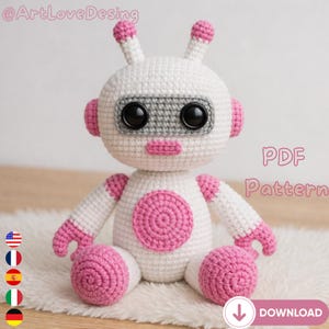 Puede incluir: Un juguete de robot de ganchillo hecho a mano en blanco y rosa. El robot tiene ojos negros grandes, una banda gris en la cara y detalles rosas en las orejas, brazos y pies. El texto "PDF Pattern" es visible.