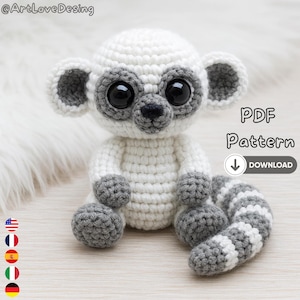 May include: A crocheted lemur toy in white and grey, with large black eyes and grey detailing on the ears, paws, and tail. The toy is sitting on a light surface. The image includes the text "PDF Pattern Download".