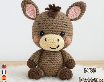 Op de afbeelding: Een handgemaakte gehaakte ezelknuffel in bruintinten, met een lichtbeige snuit en roze binnenoren. De ezel heeft zwarte ogen en roze wangen. De afbeelding bevat de tekst "PDF Pattern" en een downloadpictogram, wat duidt op een patroon om het speelgoed te maken.