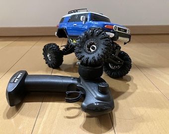 May include: A blue and black remote control toy truck with large tires. The truck is sitting on a wooden floor. The remote control is black and has a trigger.