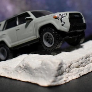 Puede incluir: Un modelo blanco de Toyota 4Runner con neumáticos y llantas negras está posicionado sobre una roca blanca texturizada. El vehículo tiene detalles en negro y una parrilla delantera negra. El fondo es borroso, azul oscuro y morado.