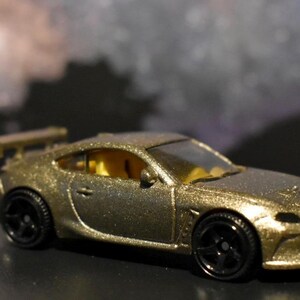 May include: A gold-colored toy car with black wheels and a rear spoiler. The miniature vehicle has a sleek design and a yellow interior, suggesting a sports car theme. The car is set against a blurred background.